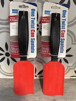 Photo of two new One Twist Can Spatulas in packaging with black handles and red spatula blades, showing 'Twist & Empty' text on packaging.