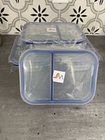 Four rectangular glass storage containers, each divided into two compartments, stacked and wrapped in plastic with lids featuring blue silicone seals and Jinamart branding.