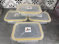 Four new rectangular glass casserole dishes stacked with yellow-rimmed lids wrapped in plastic on countertop.