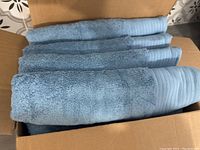 Four blue cotton bath towels folded and packed vertically inside a cardboard box, showcasing texture and stripe detail