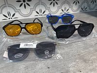 Four pairs of sunglasses displayed on a surface showing black frames with yellow lenses, black aviator style, blue wayfarer style, and black wayfarer still in packaging.