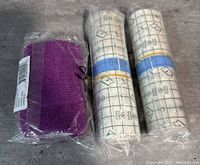 Three rolls of bandages on a gray surface: one purple cohesive bandage roll still in plastic packaging and two transparent stretch bandage rolls wrapped in plastic with grid pattern on protective cover.