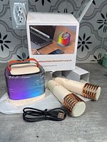 Photo of wireless karaoke speaker with rainbow LED color-changing lights, two wireless microphones with gold mesh heads, USB charging cable, and box in background.