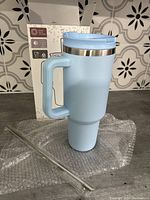 Front and side view of the tall light blue tumbler with handle, lid, and straw resting on bubble wrap, showing the packaging box behind it.