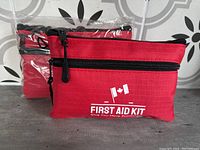 Two red first aid kit pouches, one sealed in plastic and one out, placed against a tiled background. Both have black zippers and Canadian flag with 'FIRST AID KIT' text on front.