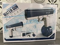 Boxed electric water gun with rechargeable continuous emission feature, showing product front and side views, packaging in white and blue with branding and promotional text.