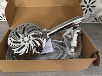 Box containing new chrome multi-spray shower head, metal hose, and bracket, showing shower head face with spray holes in a decorative pattern.