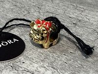 Gold-tone pig charm with red bow shown at an angle with Pandora tag and black string.