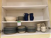 Photo of Pfaltzgraff dishware set arranged on white cabinet shelves showing bowls, plates, and mugs in cream and dark blue colors.