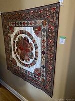 Photo of square quilted fabric tapestry hanging on wall, showing floral and geometric patchwork design in brown, beige, and maroon colors.