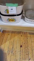 Three vintage Corningware casseroles with floral patterns and lids set on floor wood planks. Two with sunflower design, one with leafy green design.