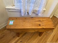 Front view of a rustic solid wood bench showing thick plank top and angled wooden legs on a hardwood floor under a window with lace curtains.