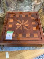 Wooden square box with decorative inlaid wood pattern on the lid, depicting a starburst design surrounded by checkerboard squares. Box is closed and placed on fabric couch.