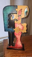 Front view showing the interlocking wooden puzzle pieces forming an abstract figure on a wooden base, colors include teal, black, beige, red, and white.