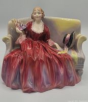 Front view of Royal Doulton figurine showing woman in red dress seated on loveseat, holding fan with hat on armrest.