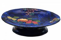 Side angled view of blue Titian Ware cake plate showing hand painted fruit decoration and pedestal base.