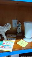 Shelf view of white elephant figurine, Elvis figure, white milk glass vase, and Rose Bowl booklets arranged on wood shelf.