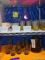 Small clear glasses, amber glasses, white Fire King ramekins, and beige mugs on a shelf.