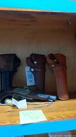 Three leather gun holsters on wooden shelf with one black powder revolver lying in front
