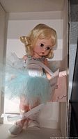 Photo showing the Madame Alexander Blue Rose Ballerina doll dressed in blue tutu and white ballet shoes with ribbon tied, lying in its original box.