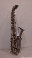 Full view of the alto saxophone standing upright against white textured wall showing the entire body and bell with a MaxSold tag attached to the bell.