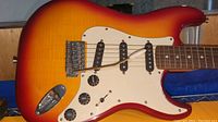 Close-up of guitar body showing sunburst finish, three single-coil pickups, white pickguard, and control knobs.