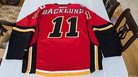 Back view of the red Reebok Calgary Flames jersey with the name 'BACKLUND' and number '11' displayed. Shows the black, yellow, and white color accents on the sleeves and lower hem.