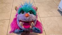 Front view of vibrant purple, pink, and blue My Pet Monster Wogster plush puppet with open mouth showing large pink tongue and visible teeth, orange nose, and green tuft on head.