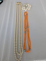 Overall view of the pearl necklace, orange multi-strand beaded necklace, and white seashell bracelet laid on fabric surface
