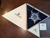 Top view of the open triangular Swarovski box containing the star snowflake ornament, certificate of authenticity, and a booklet. Box is white with Swarovski branding.