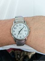 Close-up photo of Timex wristwatch on a wrist, showing white dial with black numbers, date, and silver metal expansion band.