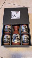Overhead view of the black gift box containing the pottery sake set: one decanter and four cups with traditional Japanese landscape designs.