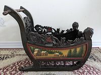 Side profile showing the dark carved wood and painted rural scenes on the sleigh panels with metal runners visible below.