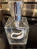 Photo showing a square clear glass lamp bergere with a silver metal top and a white wick inside on a stove surface.