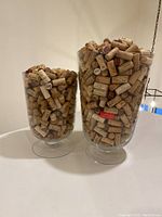 Photo showing two glass vases filled with wine corks, placed on a white surface against a plain wall.