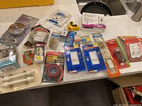 Hardware and electrical items displayed on counter including packaged doorstops, light switch covers, screws, fuse, cupboard handles, tape measure and hand tools.