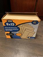 Box of 250 natural kraft open end envelopes, 9x12 size, shown closed and upright on wooden floor.