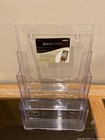 Front view of clear plastic literature holder with four tiers for business cards or brochures, showing tag inside with 'docuholder' label and mounting holes.
