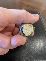 Close-up photo of the small round pendant watch being held in hand, showing blue and gold enameled exterior and white watch face with gold numbers and hands.