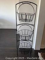 Full front view of the black metal stand with three hanging basket tiers on a dark floor, against a white wall.