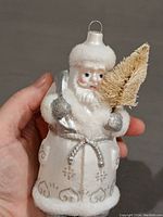 Close-up of Santa Claus ornament from the front showing frosted white coat with silver glitter detail, textured hat rim, holding golden tree.