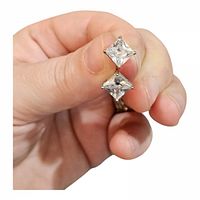 Close-up of two princess-cut stud earrings held between fingers showing clear stones in silver-tone settings.
