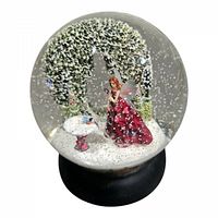 Photo showing the entire glass snow globe with fairy in red dress, green arch, and bird bath with white snow flakes inside.