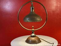 Front view of antique metal desk lamp with circular frame and ornate metal shade sitting on a white surface against red wall.