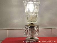 Lit Austria crystal table lamp showing clear patterned glass shade, brass floral top with crystal prisms, and white marble base.