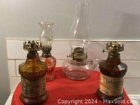 Three vintage oil lamps shown together on a red cloth backdrop. Two amber-colored lamps with metal tops and one clear glass lamp with chimney.