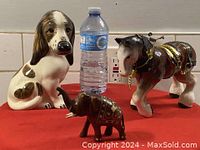 Three figurines: porcelain dog, porcelain horse with decorative harness, small wooden elephant with gold and red paint, next to water bottle for scale