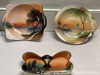 Three Noritake ceramic dishes with hand painted landscape scenes featuring trees by water under a warm sunset sky, arranged on white surface.