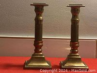 Front view of pair of solid brass candle holders showing square bases, fluted columns, and decorative details.