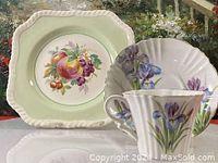 Photo showing Royal Albert teacup with purple iris flowers and matching saucer, placed in front of cake plate with fruit design.